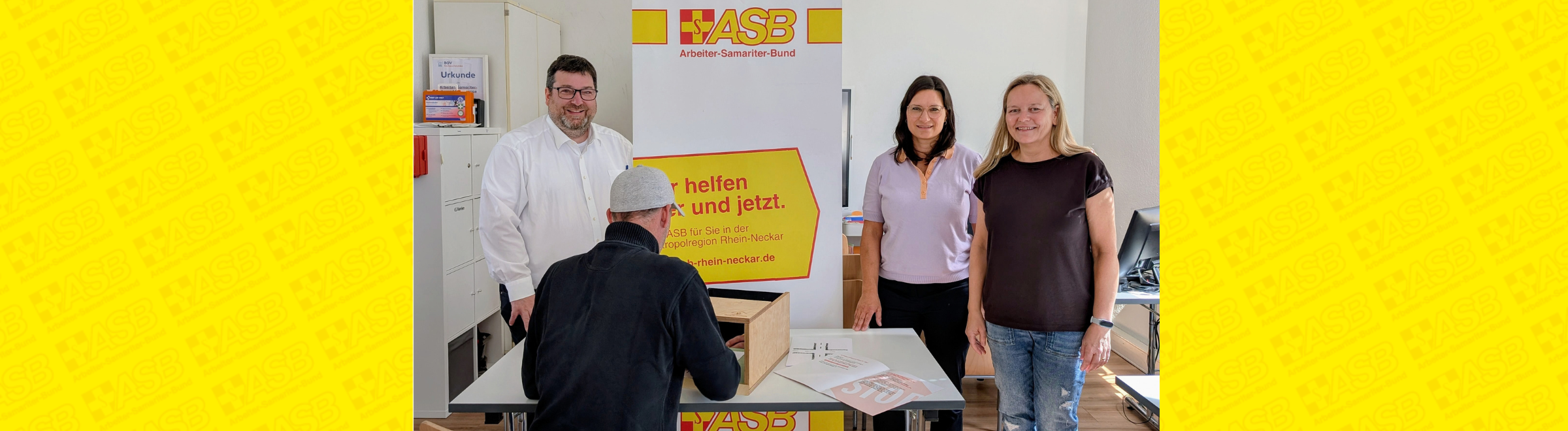 Drei Menschen stehen vor einem ASB-Roll-up und lächeln in die Kamera.