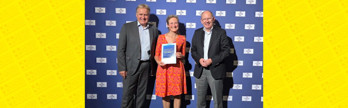 Zwei Männer und eine Frau stehen vor einer Fotowand. Die Frau hält die Auszeichnung Fuchs-Förderpreis in der Hand.