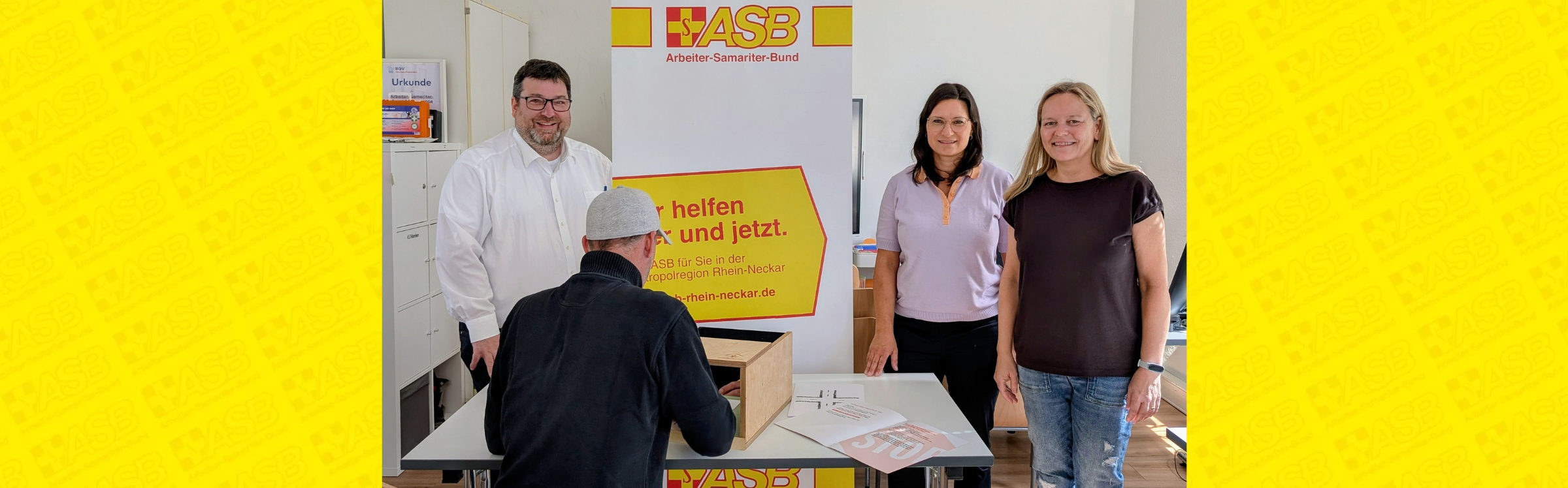 Drei Menschen stehen vor einem ASB-Roll-up und lächeln in die Kamera.